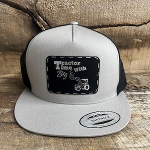 May include: A gray and black trucker hat with a curved brim. The front panel is gray with a black patch that reads "Tractor Time with Big" and features a tractor graphic. The hat has a black mesh back and a snapback closure.