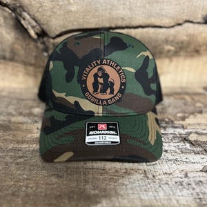 May include: A camouflage trucker hat with a brown patch featuring a gorilla and the text "Vitality Athletics Gorilla Gang". The hat is black and green with a mesh back and a Richardson 112 adjustable strap.