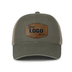 May include: An olive green baseball cap with a tan, shield-shaped leather patch centered on the front. The patch features the text "Your LOGO here" in a dark blue, stylized font with a decorative stitched border around the edge of the patch.