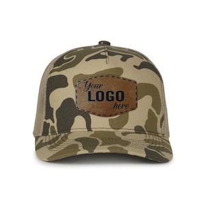 May include: Camouflage baseball cap with a brown leather patch. The patch features the text "Your LOGO here" in a dark blue font. The cap has a mesh back and a curved brim.