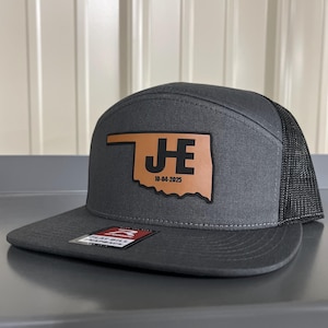 May include: A gray flat bill snapback hat with a black mesh back. The hat features a brown leather patch with the Oklahoma state outline, the initials "J E", and the date "10-04-2025".