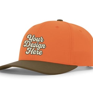May include: A bright orange baseball cap with a dark brown brim and a dark brown button on top. The front of the cap features the text "Your Design Here" in a cream-colored, cursive, retro-style font with a dark brown outline.