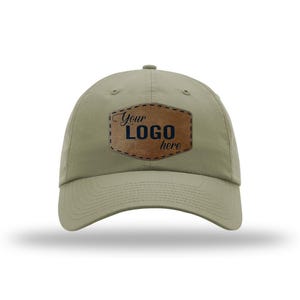 May include: A khaki-colored baseball cap with a curved brim. The cap features a brown leather patch with the words "Your Logo here" in black lettering. The hat is a neutral color and suitable for various occasions.