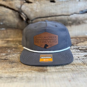 May include: A charcoal gray baseball cap featuring a white rope detail across the base of the crown. A brown hexagonal patch is centered on the front with the text GYPSUM CREEK RETRIEVERS and a dog silhouette. A rectangular sticker is on the brim.