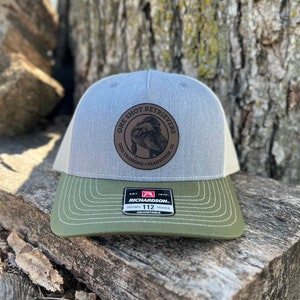 May include: A gray and green trucker hat with a brown leather patch that says "One Shot Retrievers Dog Training - Frankston, TX". The hat is made by Richardson 112.