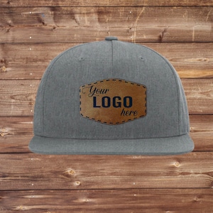 May include: A gray flat-brimmed baseball cap with a brown leather patch. The patch features the text "Your LOGO here" in a dark font. The hat has a structured crown and a flat brim.