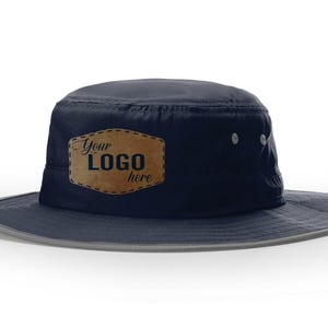 May include: A dark navy blue bucket hat featuring a tan, rectangular leather-like patch on the front with the text "Your LOGO here" printed in dark blue. The hat has a wide brim with a light grey trim and two small silver ventilation eyelets.