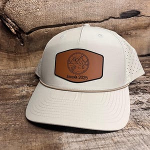 May include: A light beige baseball cap with a brown leather patch. The patch features a desert landscape design with mountains, a cactus, and the text "Sedona 2025". The cap has a curved brim and a beige rope detail.