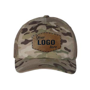 May include: A camouflage baseball cap featuring a tan, brown, and green pattern. Centered on the front is a brown, shield-shaped leather patch with black stitched edges. The text "Your LOGO here" is printed in black on the leather patch.
