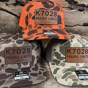 May include: Three camouflage trucker hats with the text "K7028 PRAIRIE LIMITS Outfitters" on a leather patch. One hat is orange with a black mesh back, and the others are in shades of brown and green. Each hat has a Richardson 112 tag.