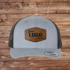 May include: Gray trucker hat with a black mesh back and a brown leather patch. The patch has the text "Your LOGO here" in a stylish font. The hat has a curved brim and a snapback closure.