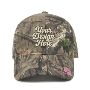 May include: A camouflage baseball cap with the text "Your Design Here" in a stylized font. The cap features a curved brim and a small, pink embroidered logo on the side. The cap is designed with a woodland camouflage pattern.