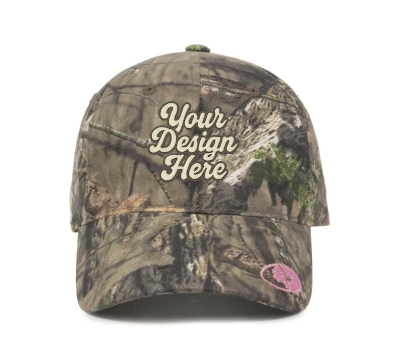 May include: A camouflage baseball cap with the text "Your Design Here" in a stylized font. The cap features a curved brim and a small, pink embroidered logo on the side. The cap is designed with a woodland camouflage pattern.