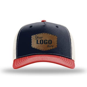 May include: A navy blue trucker hat with a red brim and white mesh back panels. The front features a brown faux leather patch with stitched edges and the text Your LOGO here in a dark font. The hat has a red button on the top center.