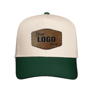 May include: A baseball cap with a cream-colored crown and a dark green brim. The cap features a brown leather patch with the text "Your LOGO here" stitched on the front. The cap is a classic style.