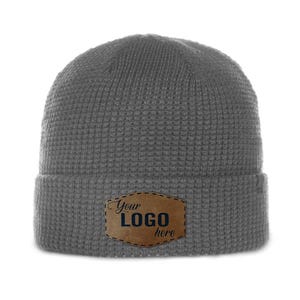 May include: A gray knit beanie with a folded brim. A brown leather patch with the text "Your LOGO here" is sewn on the front. The beanie has a textured, waffle-like pattern.