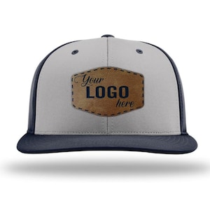 May include: A two-tone baseball cap with a light gray front panel and dark navy blue side panels and brim. Centered on the front is a brown faux leather patch with a stitched border, featuring the black text Your LOGO here.