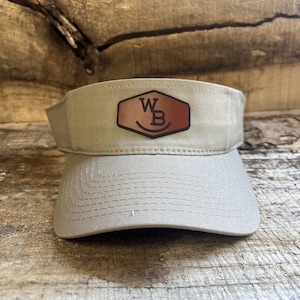 May include: A khaki-colored visor with a brown leather patch. The patch features the letters "WB" and a smiling face. The visor is made of a durable fabric and has a curved brim. The background is a wooden surface.