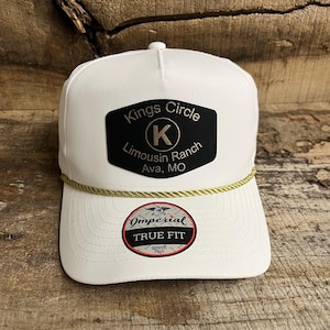 May include: White trucker hat with a black patch that reads "Kings Circle Limousin Ranch, Ava, MO" and a gold rope detail. The hat also features a "Imperial True Fit" patch.