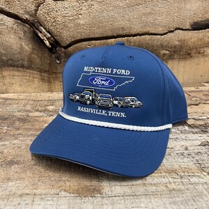 May include: A navy blue baseball cap featuring a white braided cord across the base of the crown. The front displays an embroidered graphic of the state of Tennessee, the Ford logo, and several vehicles with the text MID-TENN FORD NASHVILLE, TENN.