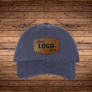 May include: A navy blue baseball cap with a brown leather patch. The patch is stitched onto the cap and features the words "Your LOGO here". The cap is made of a soft material and has a curved brim.