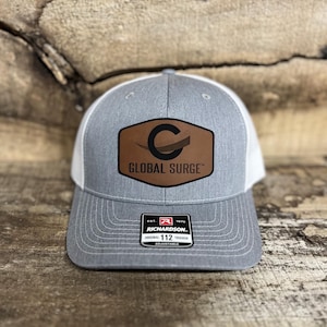 May include: A gray and white trucker hat with a brown leather patch featuring the text "GLOBAL SURGE" and a stylized "C" logo. The hat has a curved brim and an adjustable strap. The Richardson brand tag is visible.
