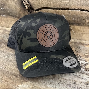 May include: A black camouflage baseball cap with a brown leather patch featuring the text "STINGRAY BLADES". The cap has a black mesh back and a snapback closure. A yellow sticker with "MULTICAM" is visible.