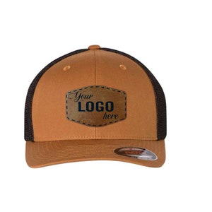 May include: A tan baseball cap with a black mesh back and a brown faux leather patch on the front. The patch features the text "Your LOGO here" in dark blue lettering. A small circular sticker is visible on the brim of the cap.
