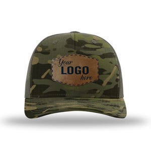 May include: A camouflage baseball cap with a brown leather patch. The patch has the text "Your LOGO here" in a stylized font. The cap features a mesh back and a curved brim.