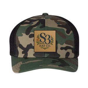 May include: A camouflage print trucker hat with a black mesh back and a tan square patch on the front. The patch features a dark blue S3 logo, a small orange flower, green leaves, and the text HAT CO. printed in dark blue capital letters.