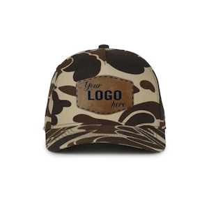 May include: A camouflage baseball cap with a brown leather patch. The patch features the text "Your LOGO here" in a stylized font. The cap has a curved brim and a button on top.