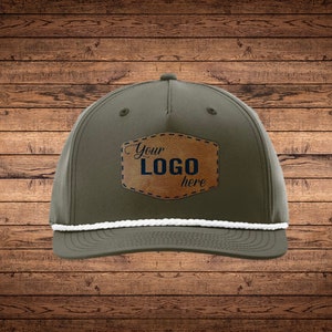 May include: Olive green cap with a white rope detail and a brown leather patch. The patch features the text "Your LOGO here" in navy blue. The cap is set against a wooden background.