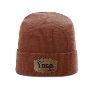 May include: A solid rust-colored knit beanie featuring a folded cuff. Centered on the cuff is a tan, shield-shaped leather-like patch with a stitched border. The patch displays the dark blue text "Your LOGO here" in a mix of script and sans-serif fonts.