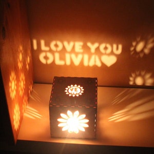 Puede incluir: Una caja de madera con una luz en su interior que proyecta las palabras "Te amo Olivia" y un corazón en una pared. La caja está decorada con un diseño floral.