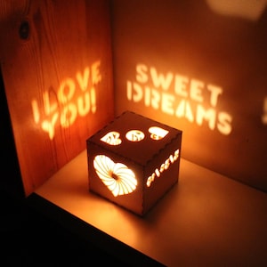 May include: A wooden box with a heart and the words "I love you!" and "Sweet Dreams" carved into it. The box is lit from inside, casting shadows of the words on the wall behind it.