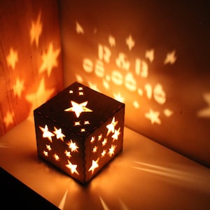 May include: A wooden cube-shaped lantern with star cutouts, casting star-shaped shadows on the wall. The lantern is illuminated from within, creating a warm glow. The date "12 & 13 05.06.16" is projected on the wall.