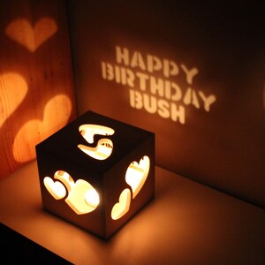 May include: A wooden box with cut-out hearts that projects the words "Happy Birthday Bush" onto a wall. The box is lit from within, casting a warm glow.