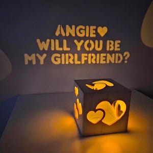 Może przedstawiać: Drewniana lampa w kształcie sześcianu z wycięciami w kształcie serc, która wyświetla na ścianie napis ANGIE SERCE WILL YOU BE MY GIRLFRIEND?. Lampa emituje ciepłe, złote światło z wnętrza, oświetlając otoczenie delikatną poświatą.
