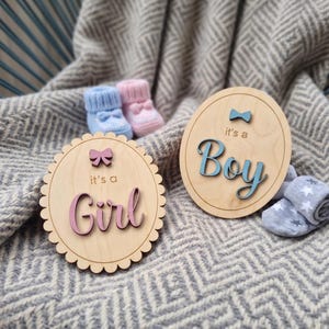 Può includere: Due targhe circolari in legno con scritte e icone in rilievo. Una targa presenta un fiocco rosa e la scritta 'it's a Girl' in rosa. L'altra targa presenta un fiocco blu e la scritta 'it's a Boy' in blu. Include scarpine da neonato blu e rosa.