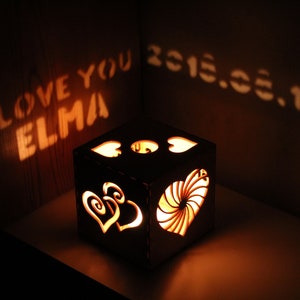 Op de afbeelding: Een houten doos met een hartvormig ontwerp en een lichtje erin. De doos werpt een schaduw op de muur die "Ik hou van je Elma 2015.03.13" zegt.