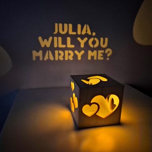 Könnte beinhalten: Ein beleuchteter Holzquader mit Herz-Ausschnitten und dem projizierten Text "JULIA, WILL YOU MARRY ME?" auf einer dunklen Oberfläche. Der Quader strahlt ein warmes, einladendes Licht aus.