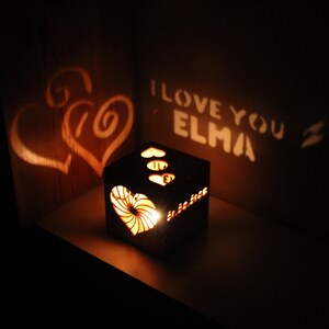 Op de afbeelding: Een houten doos met een hartvormige uitsparing die een schaduw van twee harten en de tekst "I love you Elma" op een muur projecteert. De doos wordt van binnenuit verlicht, waardoor een warme gloed ontstaat.