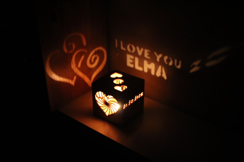 Op de afbeelding: Een houten doos met een hartvormige uitsparing die een schaduw van twee harten en de tekst "I love you Elma" op een muur projecteert. De doos wordt van binnenuit verlicht, waardoor een warme gloed ontstaat.