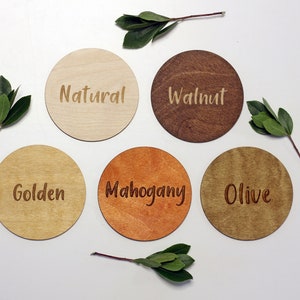 May include: Five round wooden coasters in different wood stains. The coasters are labeled "Natural", "Walnut", "Golden", "Mahogany", and "Olive".