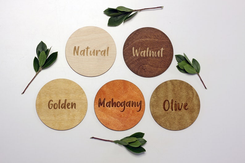 May include: Five round wooden coasters in different wood stains. The coasters are labeled "Natural", "Walnut", "Golden", "Mahogany", and "Olive".