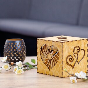 Op de afbeelding: Een houten doos met een hartvormig uitsnijding. De doos is gemaakt van lichtbruin hout en heeft een natuurlijke houtfinish. De hartvormige uitsnijding bevindt zich in het midden van de doos en is omgeven door een swirl-ontwerp. De doos staat op een houten tafel.