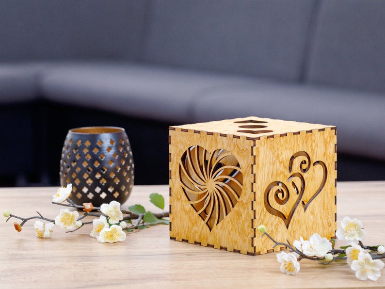 Op de afbeelding: Een houten doos met een hartvormig uitsnijding. De doos is gemaakt van lichtbruin hout en heeft een natuurlijke houtfinish. De hartvormige uitsnijding bevindt zich in het midden van de doos en is omgeven door een swirl-ontwerp. De doos staat op een houten tafel.