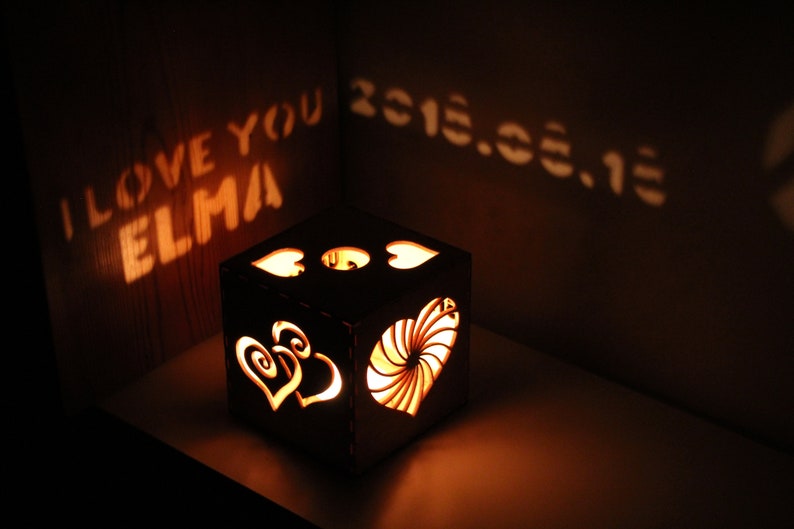 Op de afbeelding: Een houten doos met een hartvormig ontwerp en een lichtje erin. De doos werpt een schaduw op de muur die "Ik hou van je Elma 2015.03.13" zegt.