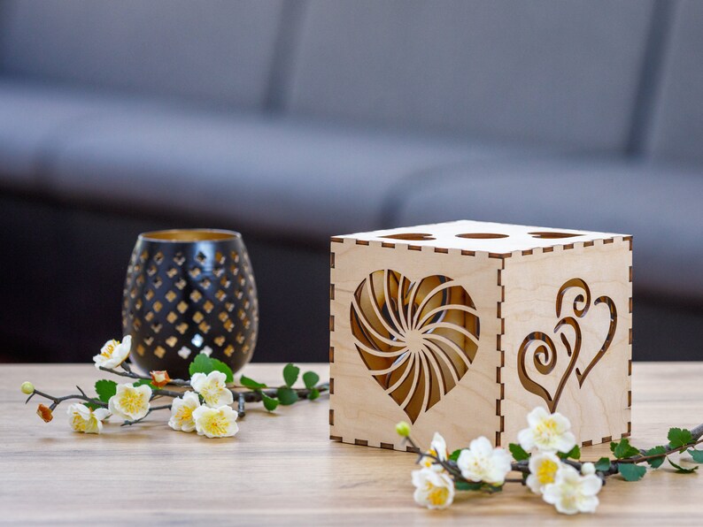 Op de afbeelding: Een houten doos met een hartvormige uitsparing aan elke kant. De doos is lichtbruin en heeft een natuurlijke houten afwerking. De hartvormige uitsparingen zijn ingewikkeld en gedetailleerd. De doos staat op een houten tafel met een klein boeket witte bloemen ervoor.