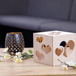 May include: A wooden box with heart-shaped cutouts. The box is light brown and has a natural wood finish. The box is sitting on a wooden table with a branch of white flowers.